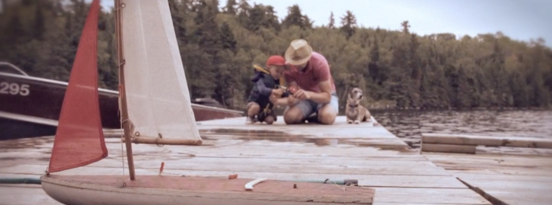 A Man and his child and dog are on a river/lake dock, attempting to fix a tool together. A toy boat is in focus at the front.