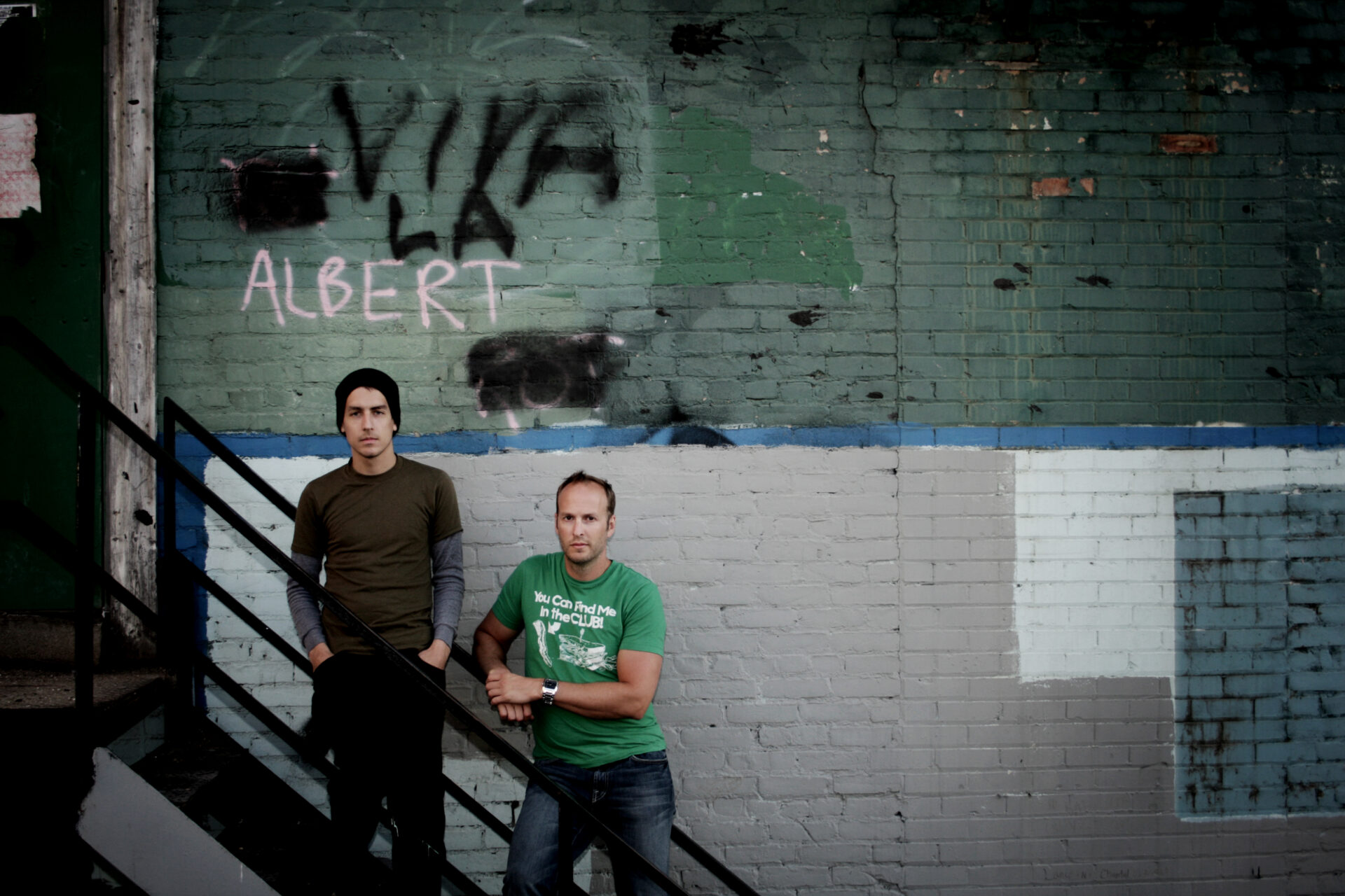Jeff Newman and Randy Frykas stand on a back-alley stairway, overtop of them is spray paint graffiti that reads "Viva La Albert"
