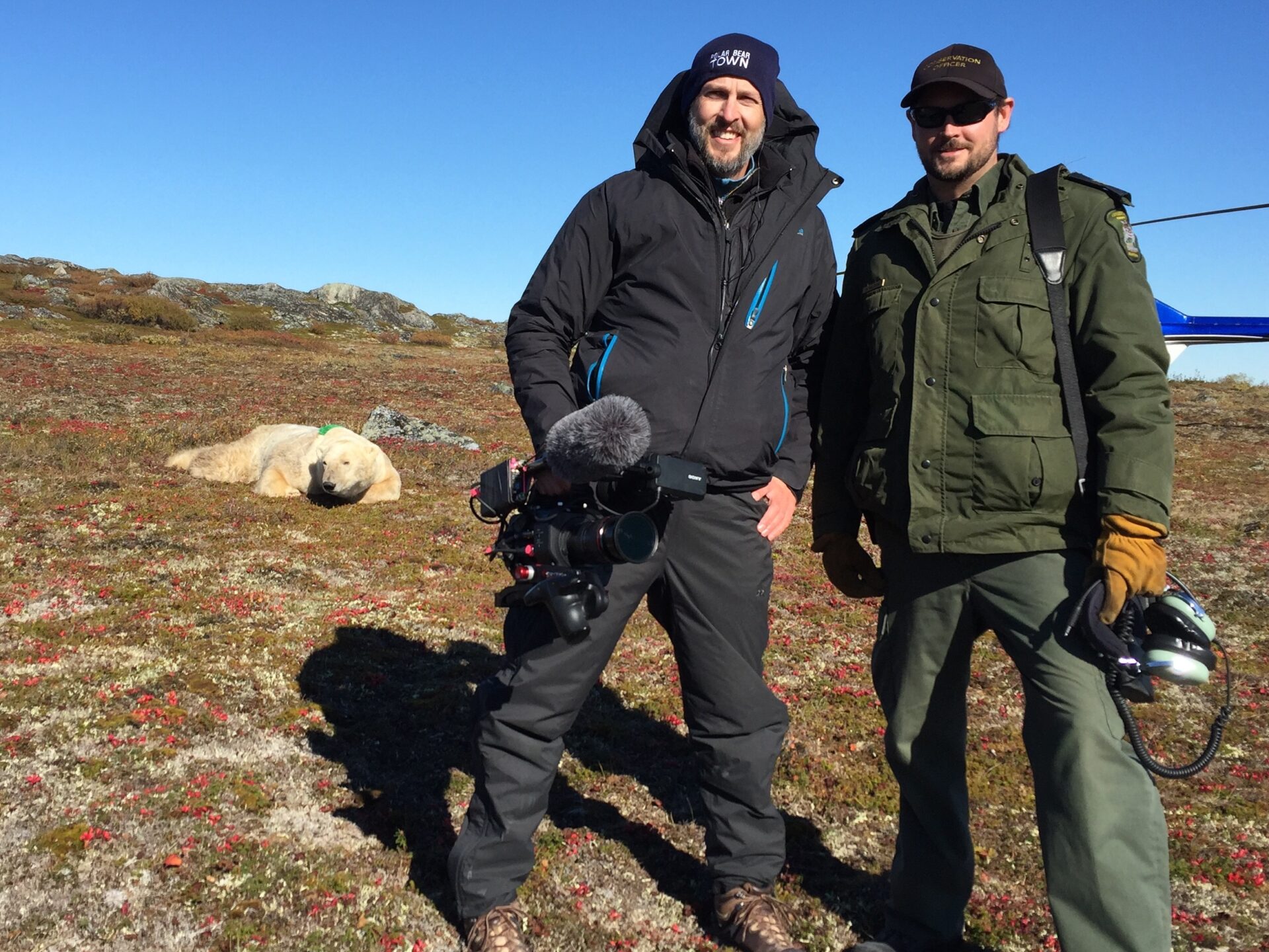 Jeff Newman and a park ranger stand smiling together with a sleepy polar bear in the background.